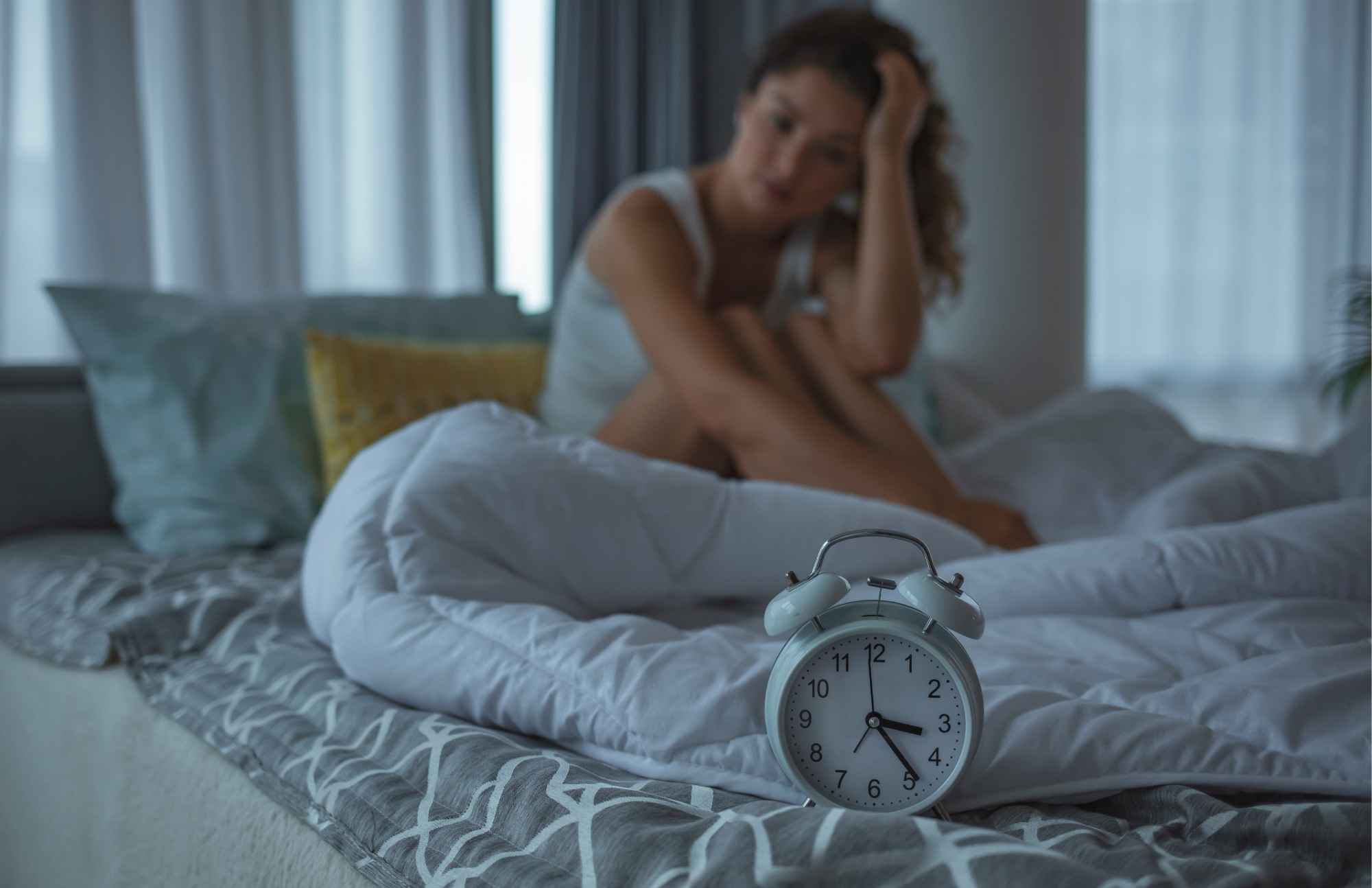 Woman struggling to sleep, sitting on a bed next to an alarm clock, representing insomnia and poor sleep due to hormone imbalance.