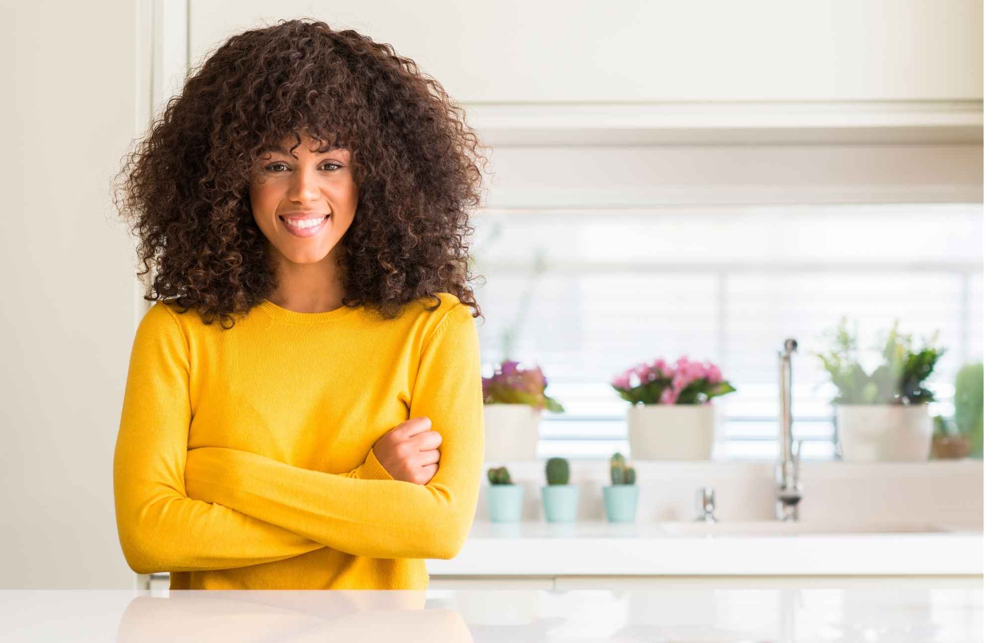 A confident woman wearing a yellow sweater, standing in a bright kitchen, representing overall wellness and proactive health management.