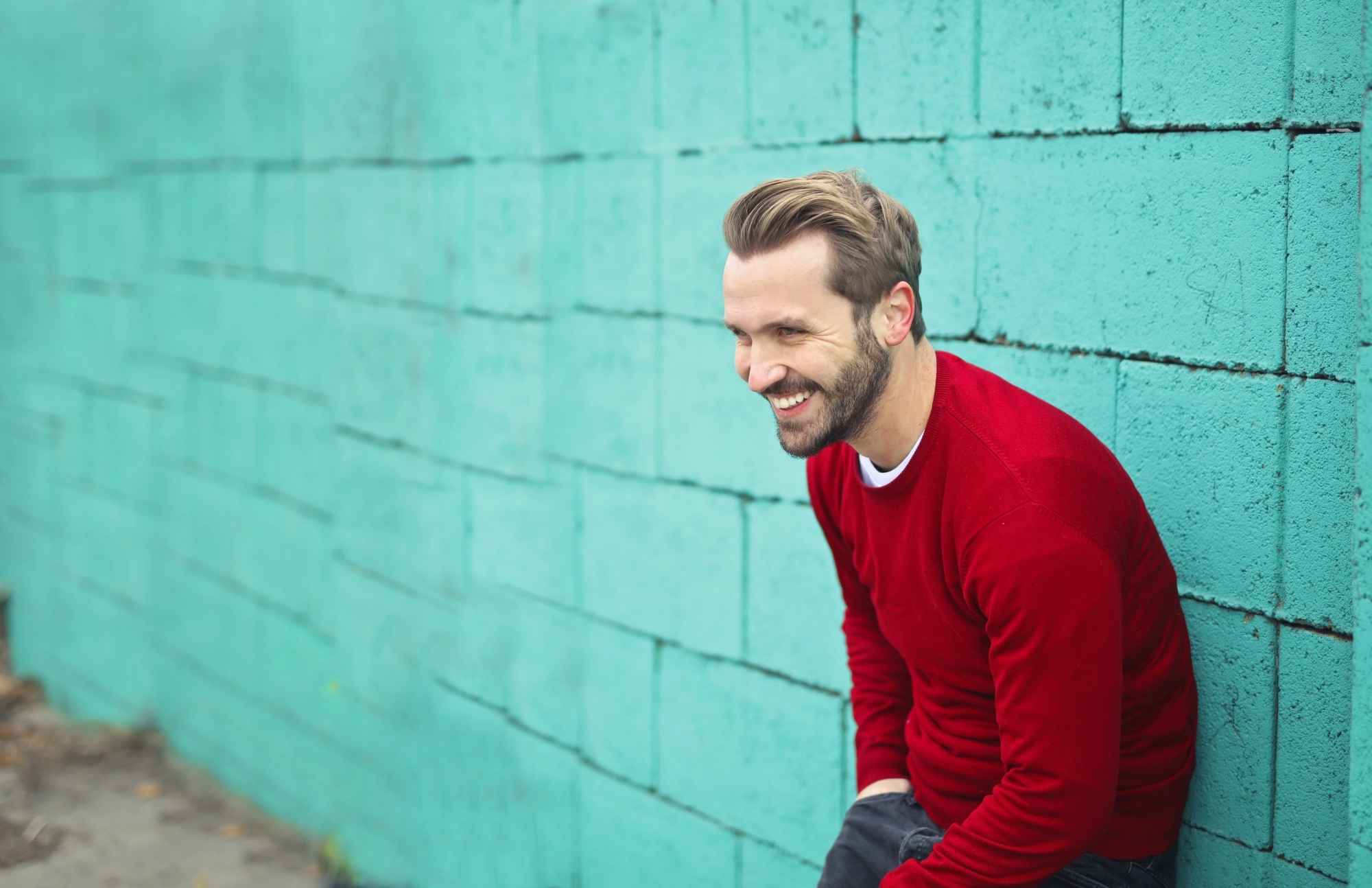 A smiling man in a red sweater sitting against a turquoise wall, symbolizing good health and well-being from routine health screenings.