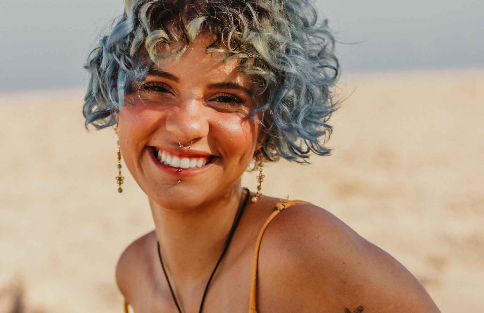 A woman with blue curly hair smiling at the beach, representing the importance of regular health checkups for maintaining vitality and happiness.