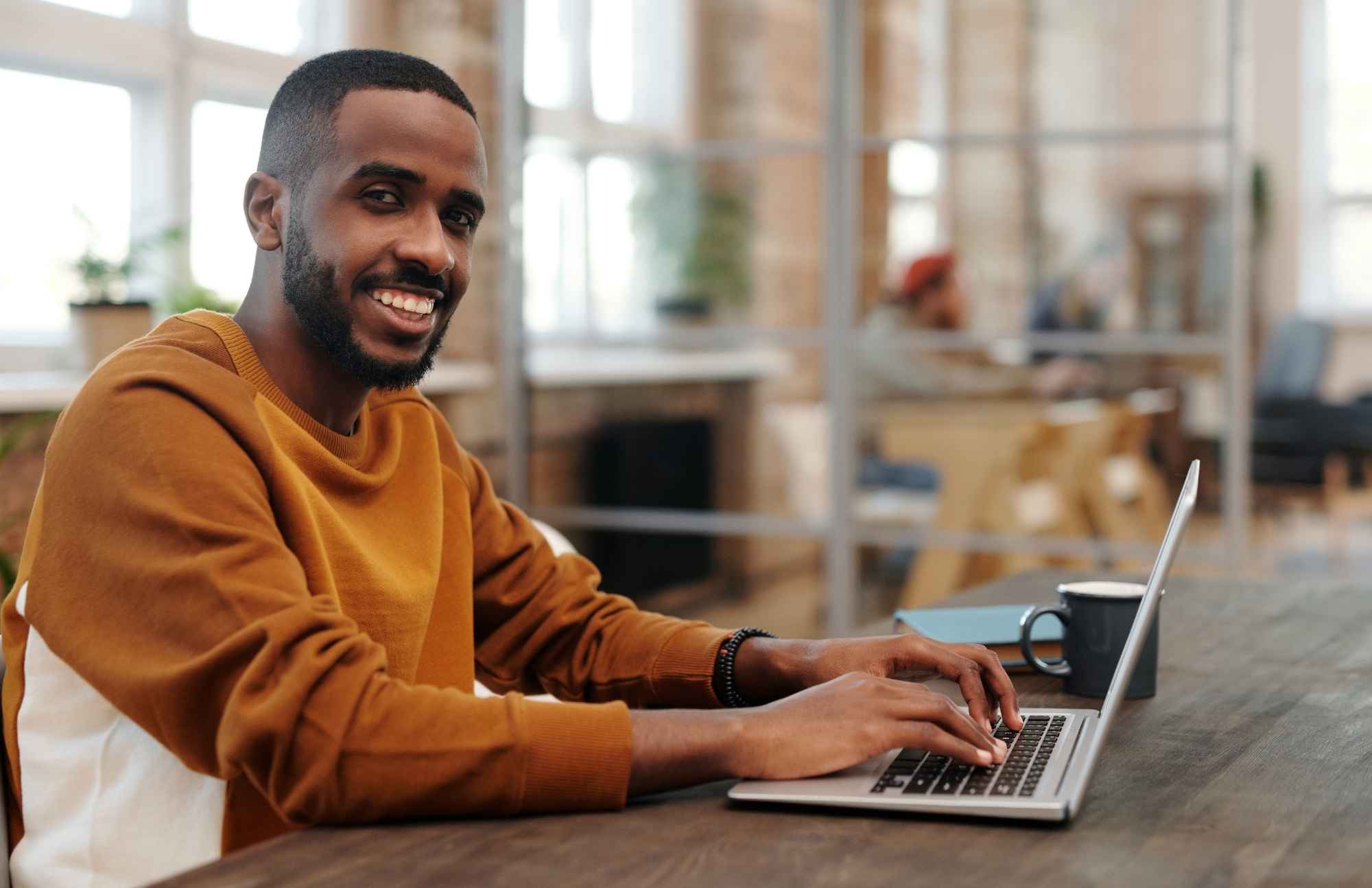 A professional man smiling while working on a laptop in a modern office, emphasizing the role of health screenings in maintaining productivity and overall wellness.