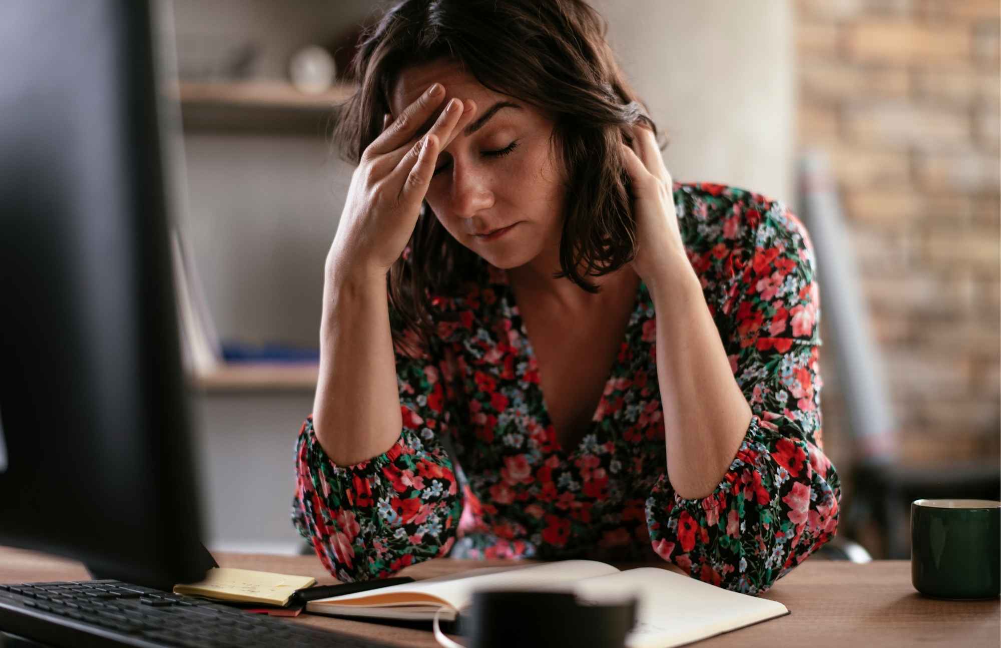 A woman sitting at a desk with a hand on her forehead, looking fatigued and in discomfort.